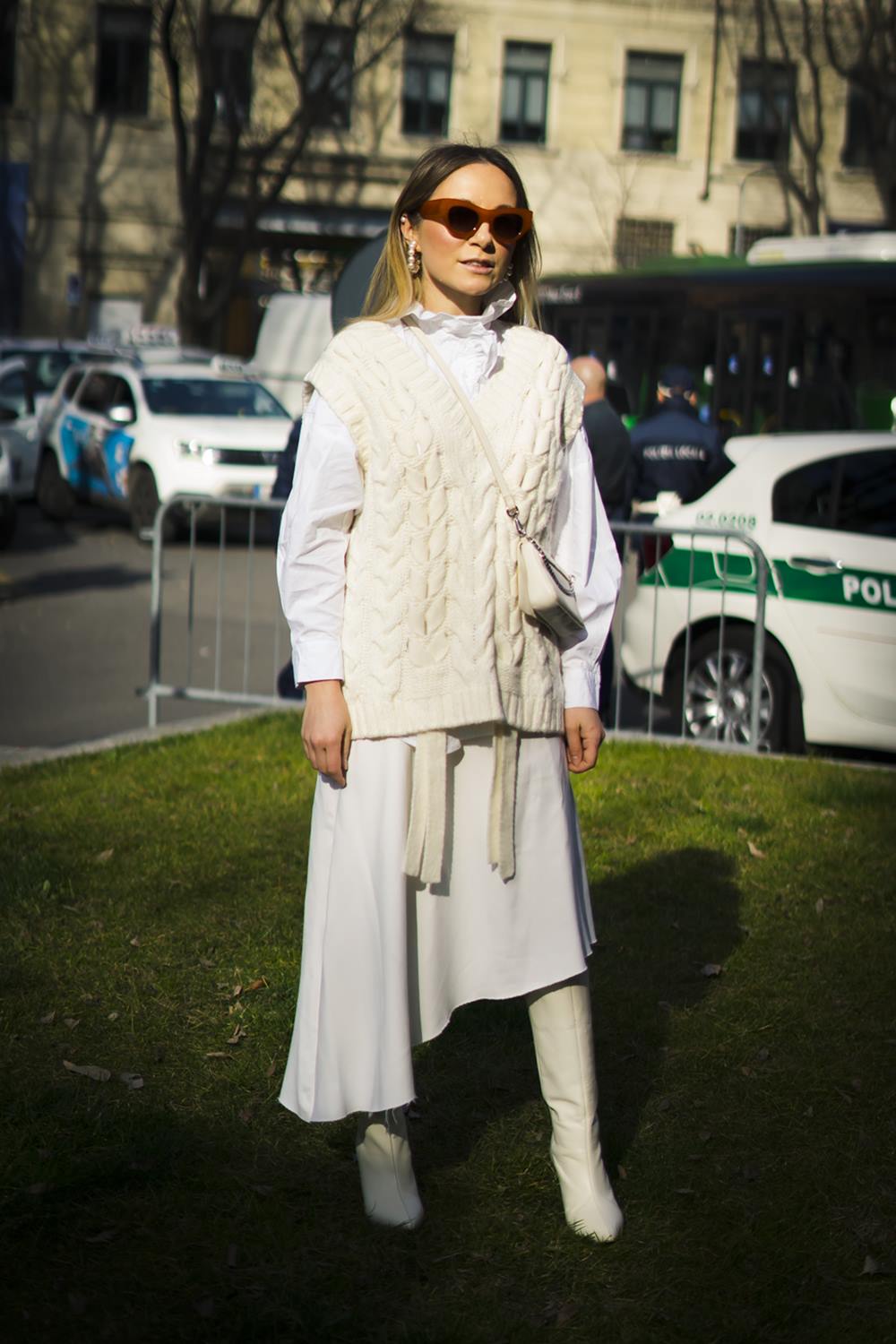 Mulher branca e jovem, de cabelo liso loiro, anda pelas ruas de Paris. Ela usa uma camisa branca, um colete branco de tricô, saia branca e bota de couro. - Metrópoles
