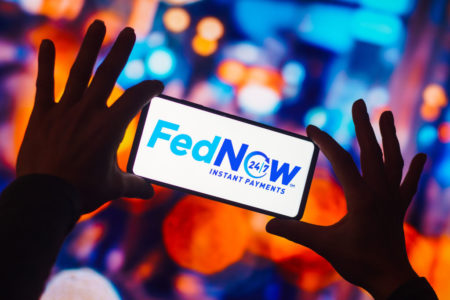 Imagem de um celular sendo segurado e exibindo na tela o logotipo do FedNow, novo sistema de pagamento dos Estados Unidos, semelhante ao Pix. Ao fundo, muitas luzes coloridas.