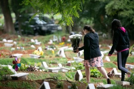 Imagem de uma mulher de paletó preto e saia rosa carregando flores em um cemitério com covas abertas e lápides brancas - Metrópoles