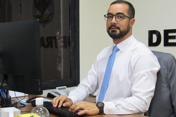 Fotografia colorida de homem de camisa branca e grava azul sentado em mesa digitando em teclado