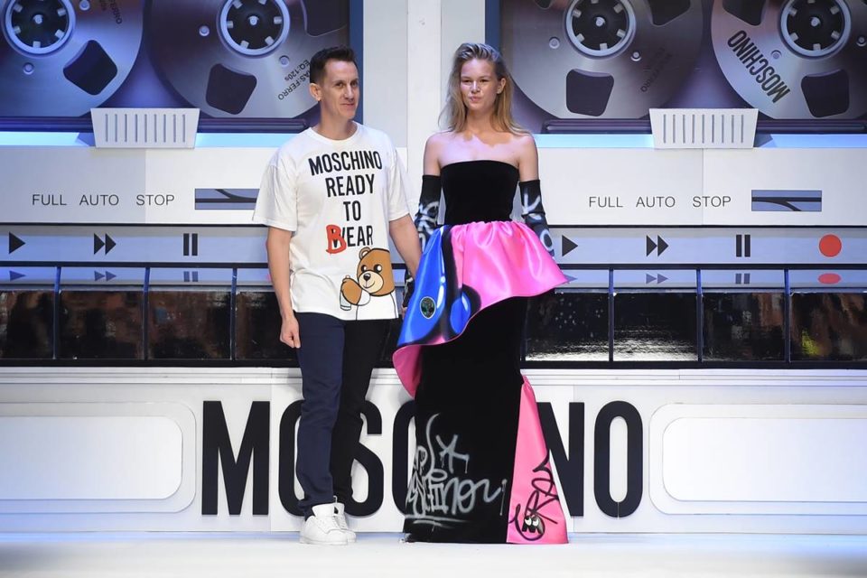 Estilista Jeremy Scott na passarela da Moschino. Ele usa camiseta branca com escrita e calça jeans, ao lado de modelo com vestido preto com detalhes em rosa e azul - Metrópoles