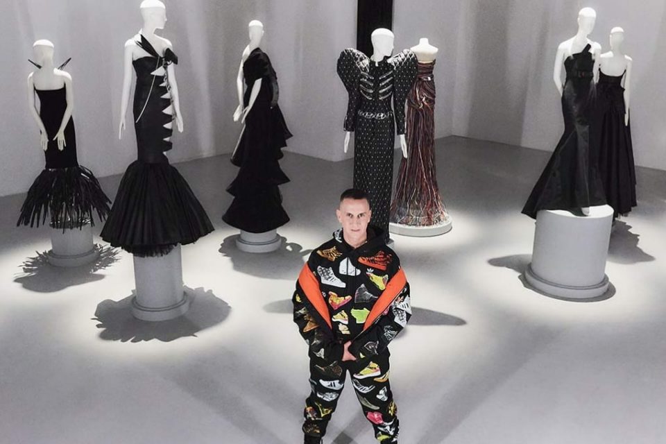 O estilista Jeremy Scott, um homem branco e de meia idade, com cabelo preto curto, posa ao lado de manequins. As modelos vestem roupas feitas por ele, vestidos pretos construídos a partir de resíduos de carros. - Metrópoles