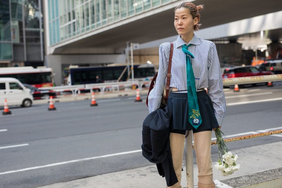 Na rua, mulher usa camisa listrada com gravata verde e saia curta azul - Metrópoles