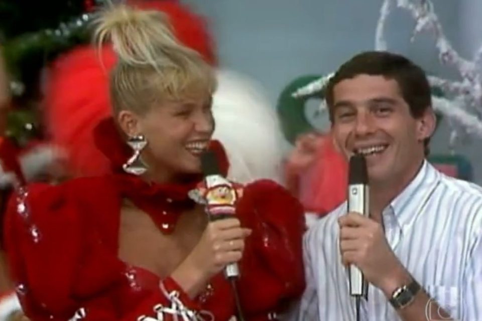 foto colorida de xuxa jovem com roupa vermelha segurando microfone e ayrton senna segurando microfone ao lado dela - metrópoles