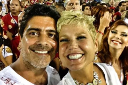 Foto colorida de Xuxa e Junno Andrade com blusas branca e sorrindo. Ao fundo, fãs vestidos de vermelho - Metrópoles