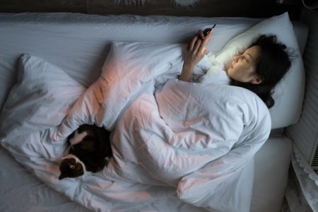 Mulher deitada na cama mexendo no celular. Um gato está deitado nos pés dela - Metrópoles