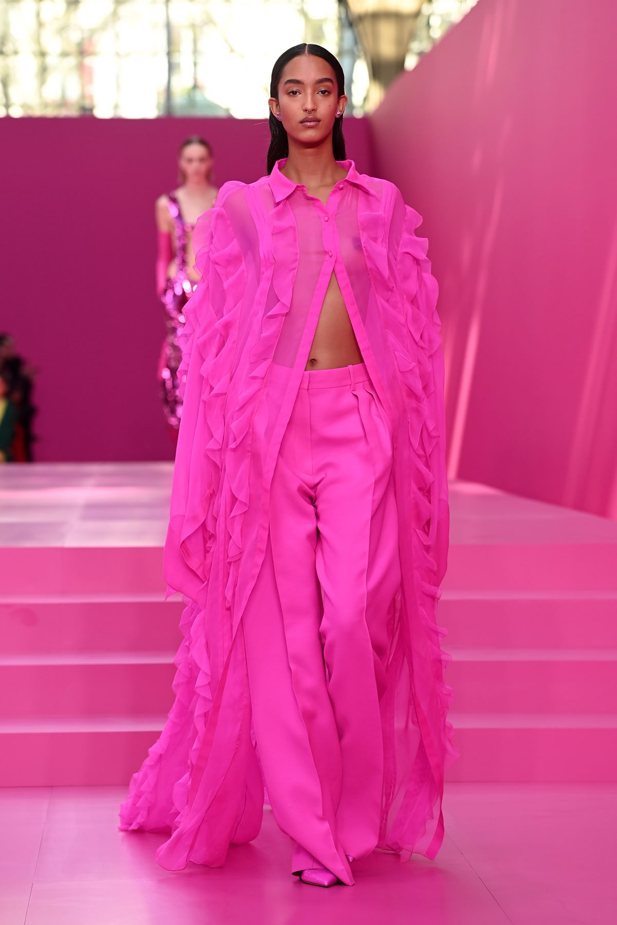 Mulher negra e jovem, de preto liso, desfila para a marca Valentino Semana de Moda de Paris. Ela usa um vestido longo e calça, tudo na cor rosa. - Metrópoles