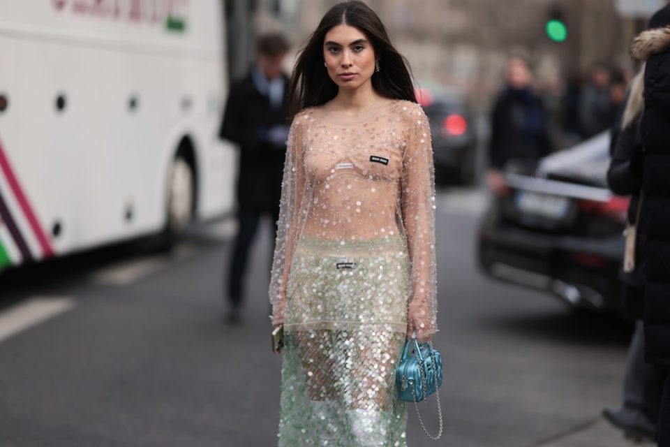 Mulher branca e jovem, de cabelo liso loiro, posa para foto na rua durante a Semana de Moda de Paris. Ela usa um sutiã bege, saia verde e, por cima, um vestido transparente com paetês. As peças são da marca Miu Miu. - Metrópoles