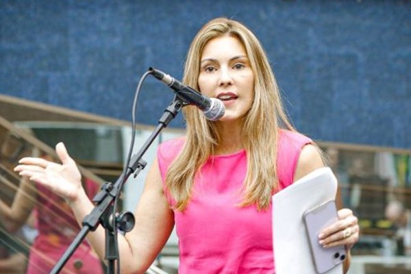 Fotografia colorida de mulher vestida de rosa falando em microfone enquanto segura celular e papeis com a mão esquerda