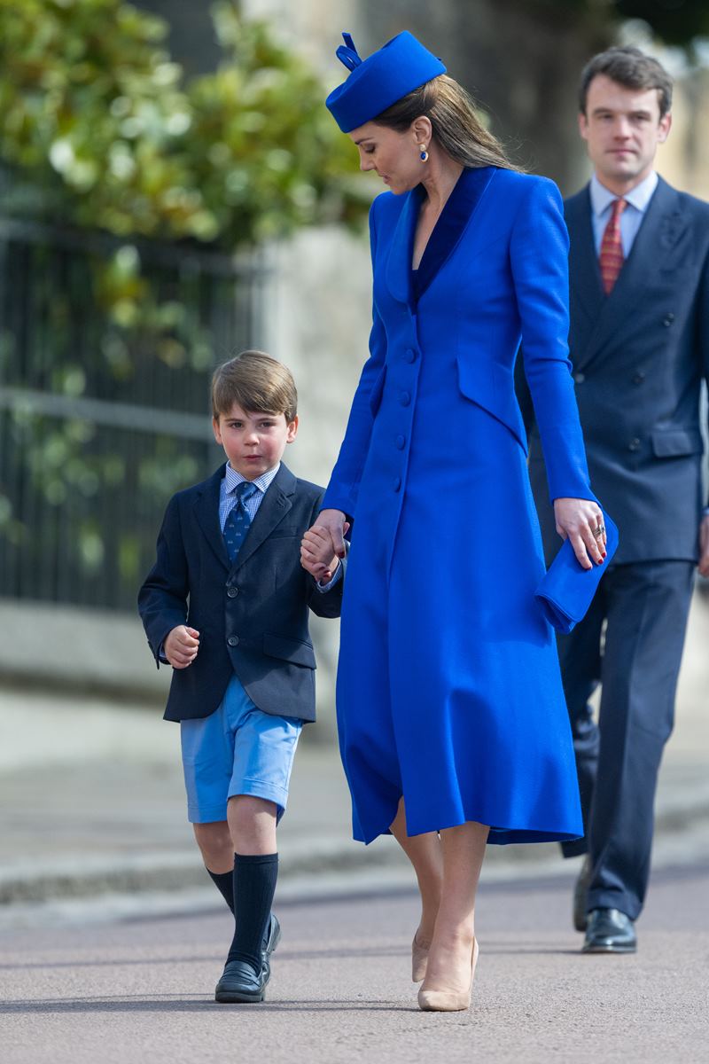 Foto colorida de menino branco, loiro, de blazer, gravata e shorts e mãos dadas com mulher branca, de cabelo castanho de mãos dadas com mulher alta, branca e de vestido na cor azul royal - Metrópoles