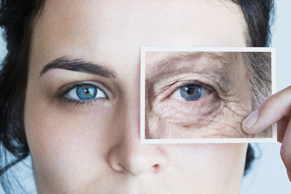 Foto colorida de mulher branca, de olhos azuis, e pele jovem. Ela segura na altura do olho uma fotografia de uma pele na região dos olhos com rugas e linhas de expressão - Metrópoles