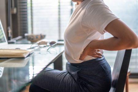 mulher com as mãos nas costas sentada em cadeira de frente para uma mesa com computador - Metrópoles