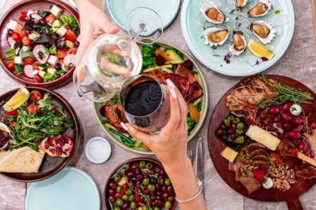 Mesa ista de cima com várias comidas e duas pessoas brindando - Metrópoles