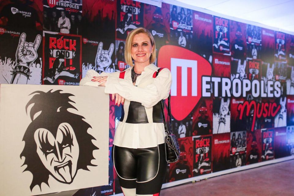 Mulher loira apoiada com os braços em caixas. Ela está posando para foto em frente a fundo com propaganda - Metrópoles