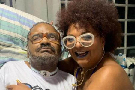 Foto colorida de Arlindo Cruz e Babi Cruz. Ele está de camisa branca e usa um aparato no pescoço. Ela está óculos branco e colar - Metrópoles