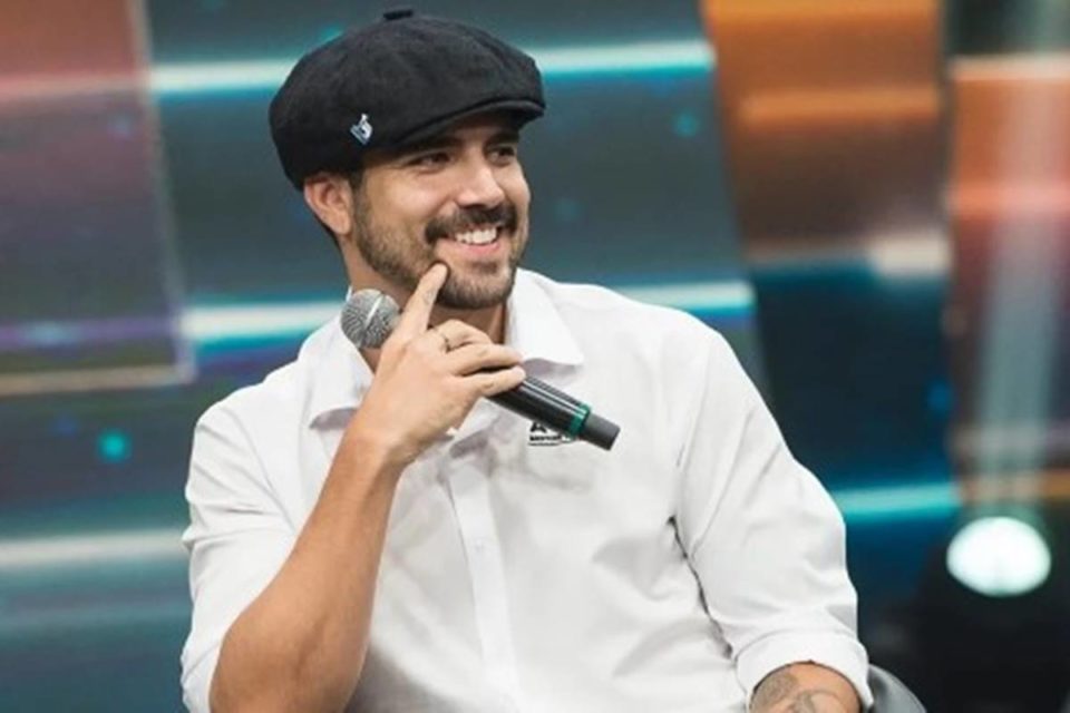 Imagem colorida do ator Caio Castro, usando boina e blusa branca, com microfone na mão - Metrópoles