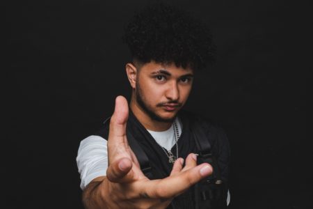 DJ Klap. Ele é um homem branco, com cabelo e barba preto. Usa um colete preto, uma camisa branca e está com a mão para a frente. O fundo é preto - Metrópoles