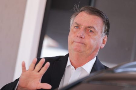 Foto colorida do Ex-presidente Jair Bolsonaro, de terno, com a mão para cima