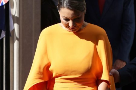 Foto colorida de mulher com um vestido laranja e uma bolsa nas mãos. Ela sai de um portão - Metrópoles