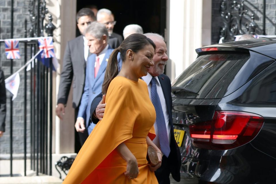 Foto colorida de mulher com um vestido laranja e uma bolsa nas mãos. Ela está ao lado de um homem branco idoso, vestido com terno - Metrópoles