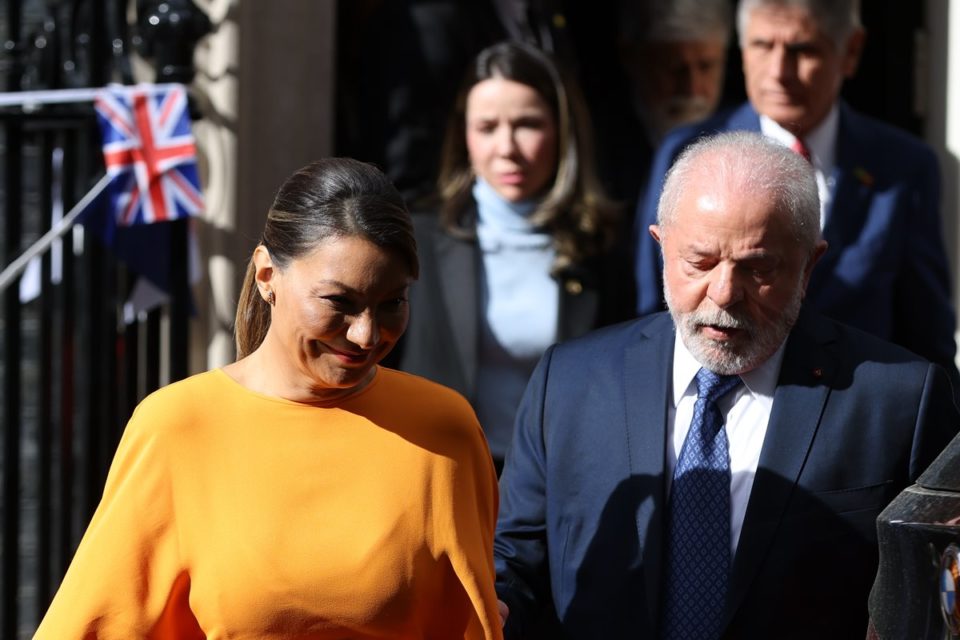 Foto colorida de mulher com um vestido laranja e uma bolsa nas mãos. Ela está ao lado de um homem branco idoso, vestido com terno - Metrópoles