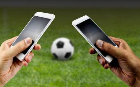 Fotografia colorida mostrando duas mãos mexendo em celulares e uma bola de futebol entre elas-Metrópoles