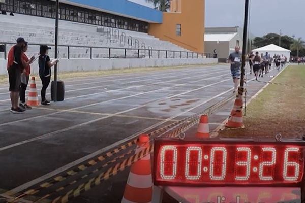 Fotografia colorida mostra cronômetro com pessoas correndo em pista de atletismo ao fundo