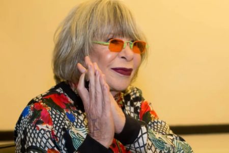 Foto colorida de Rita Lee. Ela está com cabelo grisalho, óculos laranja, batom vinho e com a palma das mãos juntas, próximas ao rosto - Metrópoles