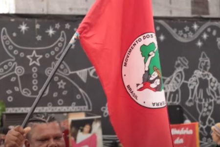 Imagem mostra bandeira do MST. A bandeira, que tem estampa em vermelho e branco, está em um local com diversos desenhos e é segurada em uma haste preta - Metrópoles