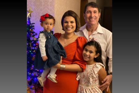 Imagem colorida: família com mãe, pai e duas filhas crianças em frente a árvore de Natal - Metrópoles