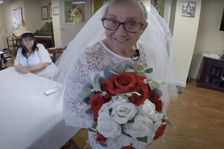 Foto colorida de uma idosa com roupa de noiva e com buque com flores vermelhas e brancas - Metrópoles