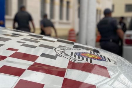 Imagem colorida do capô de uma viatura da Polícia Militar de SP. A peça é quadriculada em branco e vermelho com o logo da PM paulista. Ao fundo, alguns PMs - Metrópoles