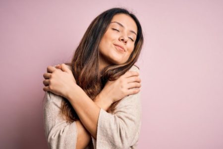 Mulher se abraçando de camisa bege e com os olhos fechados -Metrópoles