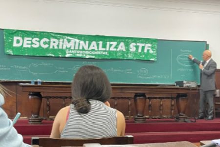 Foto colorida do ministro Alexandre de Moraes em sala de aula, na USP, com faixa de protesto: descriminaliza, STF