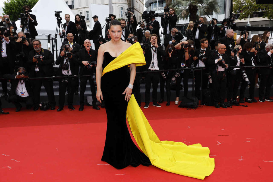 Na imagem com cor, uma mulher branca de cabelos pretos preso usando um vestido preto com um detalhe em amarelo posando em um tapete vermelho - Metrópoles