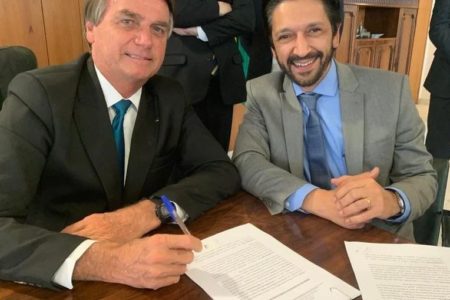 Imagem colorida mostra Ricardo Nunes, homem branco, de cabelo e barba pretos, terno cinza e gravata azul, à esquerda, e Jair Bolsonaro, homem branco, grisalho, de terno preto. Eles estão sentados em uma mesa sorrindo para câmera - Metrópoles