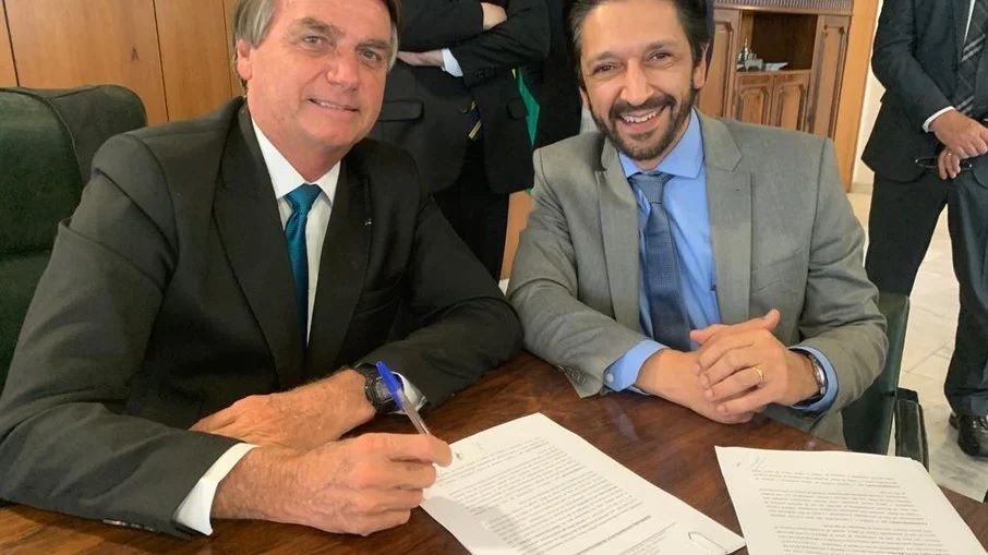 Imagem colorida mostra Ricardo Nunes, homem branco, de cabelo e barba pretos, terno cinza e gravata azul, à esquerda, e Jair Bolsonaro, homem branco, grisalho, de terno preto. Eles estão sentados em uma mesa sorrindo para câmera - Metrópoles