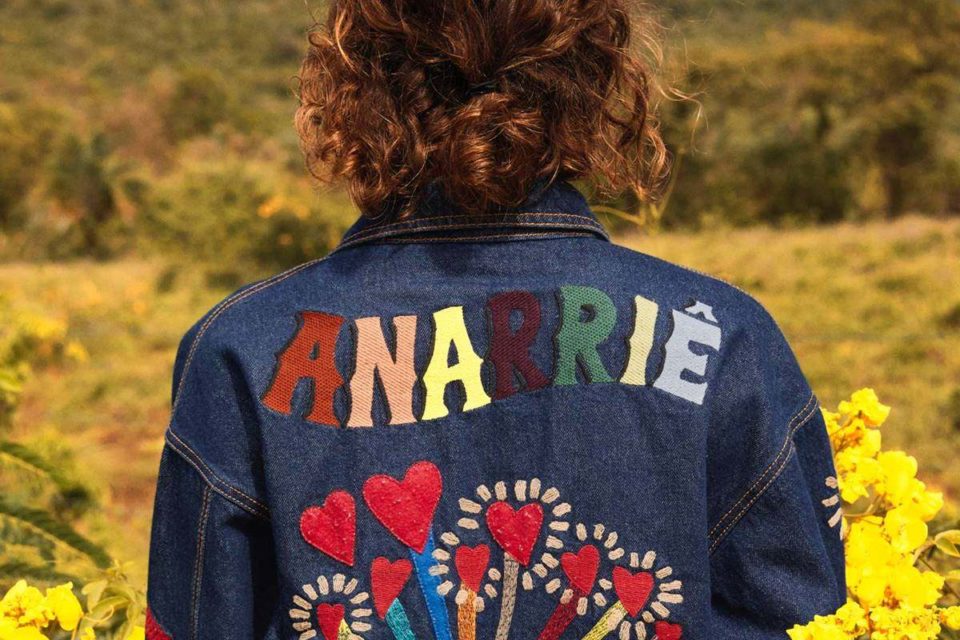 Modelo de costas usando jaqueta jeans. A peça está estampada com frase colorida - Metrópoles