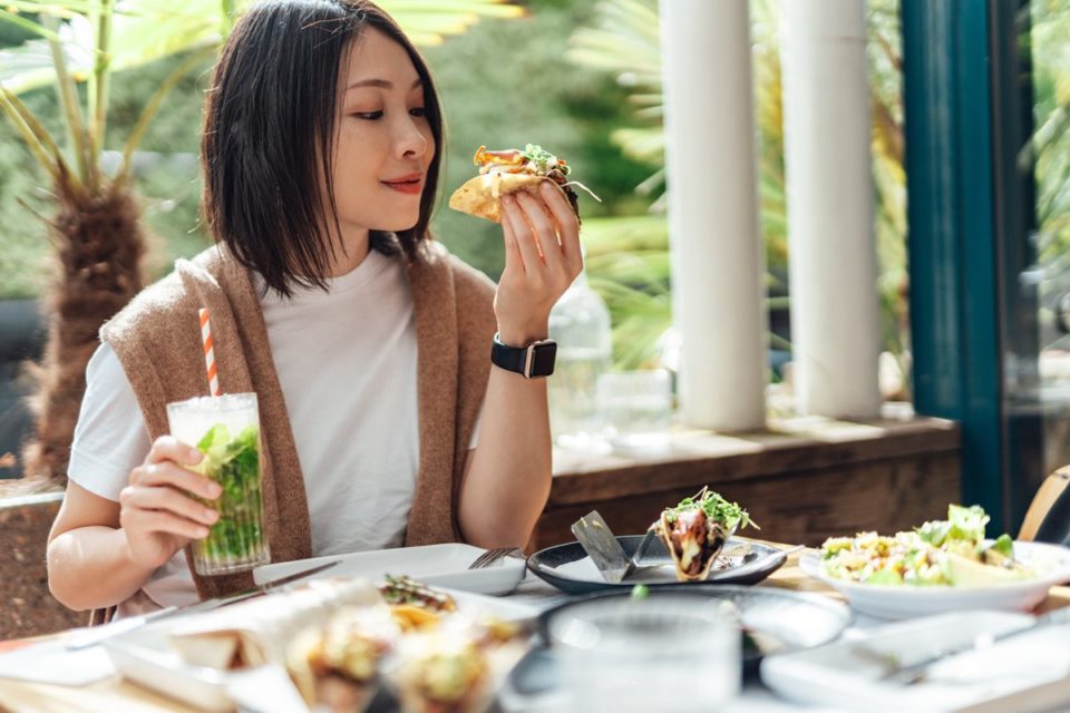 Mulher oriental segurando um copo com suco verde e um quitute na outra mão - Metrópoles