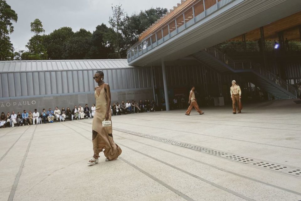 Em desfile, mulher negra usa vestido acetinado marfim com recortes. Ao fundo, é possível ver outros dois modelos, além da plateia, posicionada em volta - Metrópoles