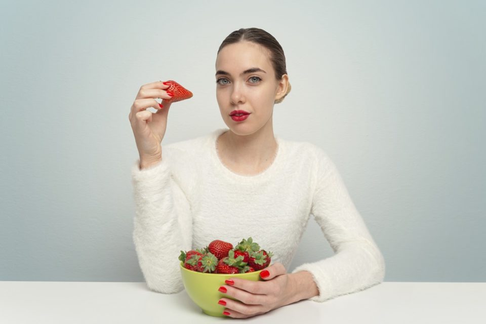 Foto colorida de mulher branca segurando um morango e uma tigela com a fruta - Metrópoles