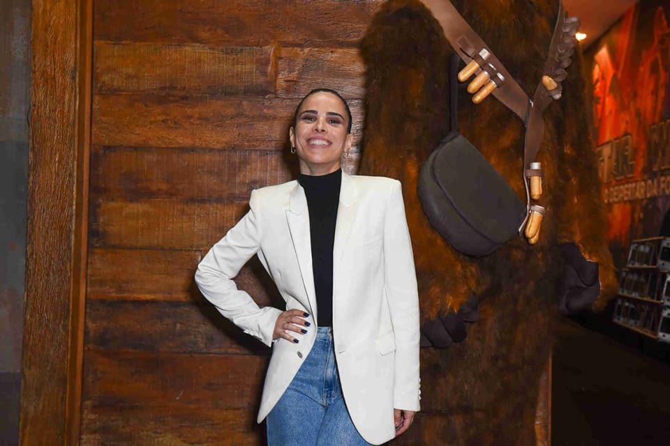 foto colorida de wanessa camargo de jeans, blusa preta e blazer branco posando com mao da cintura em fundo rustico - metrópoles