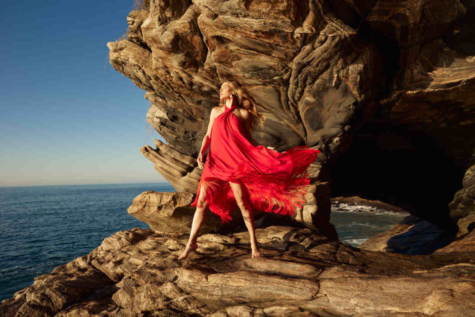 Na imagem com cor, uma mulher branca loira em uma rocha com um vestido vermelho - Metrópoles