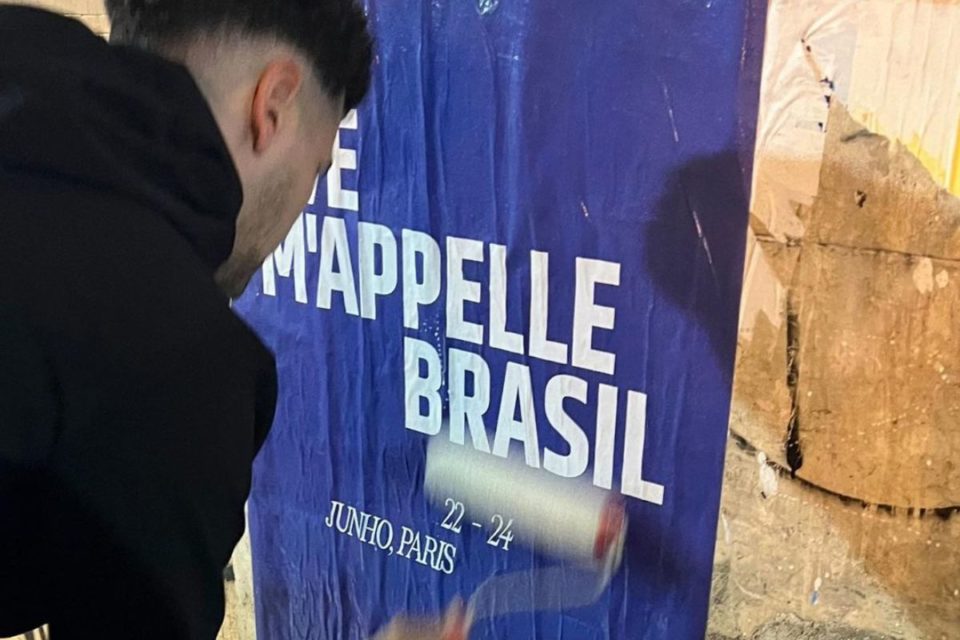 Na imagem com cor, um homem de touca colando um lambe-lambe na parede