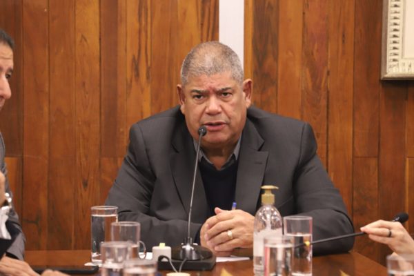 Imagem colorida mostra o vereador Milton Leite, homem negro, grisalho, de terno cinza e camisa preta, sentado em uma mesa de reuniões com microfone em uma sala de paredes de madeira - Metrópoles