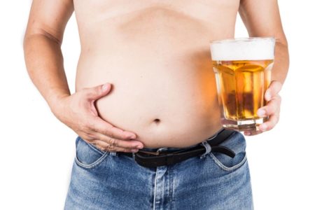 Imagem colorida de homem sem camisa, de calça jeans e a segurar copo de cerveja com a mão esquerda emquanto segura barriga com a outra