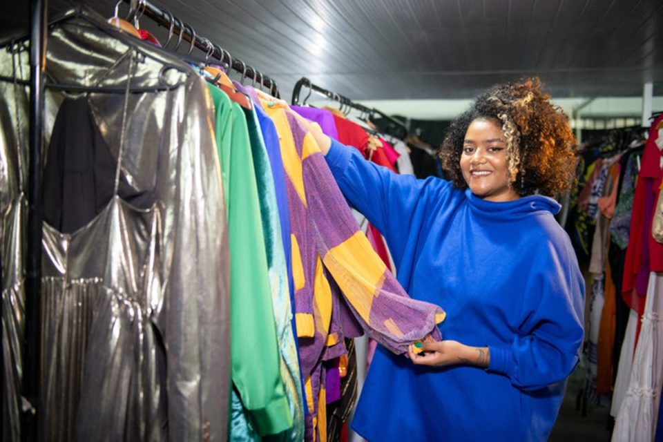 Mulher posa ao lado de arara com roupas coloridas. Ela segura a manga de um casaco roxo e amarelo - Metrópoles