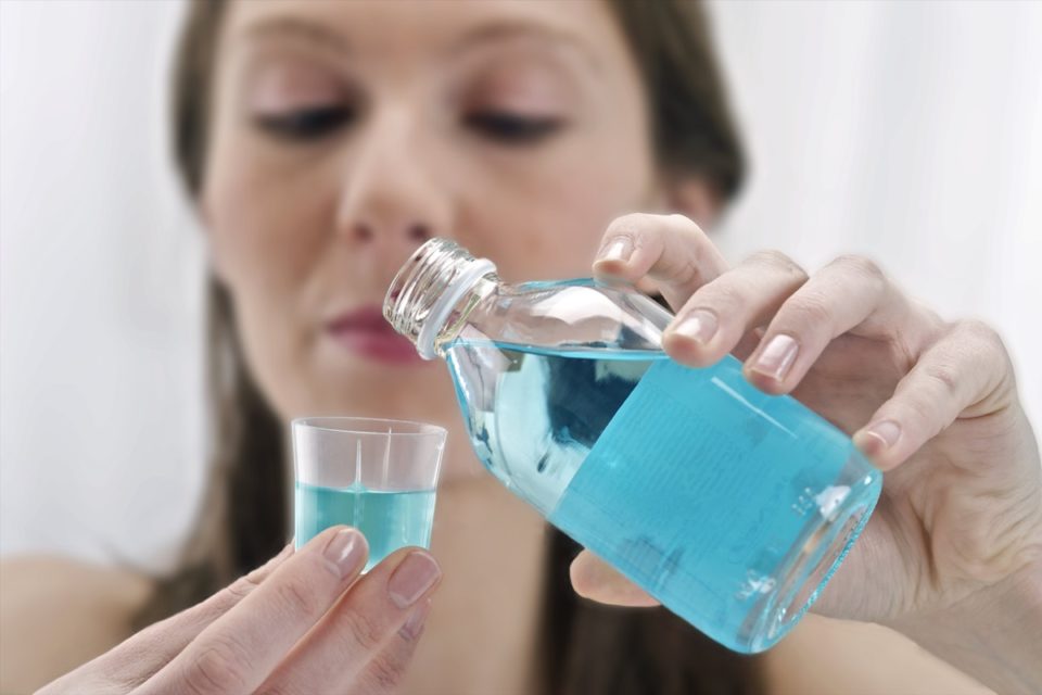 Foto colorida de mulher branca colocando enxaguante bucal dentro de um copo transparente - Metrópoles