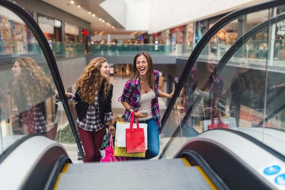 Em escada rolante, duas amigas seguram sacolas de compras - Metrópoles