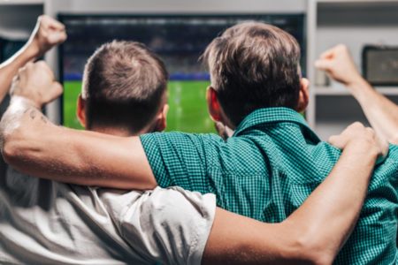 Fotografia colorida mostrando dois homens abraçados comemorando em frente à televisão-APOSTAS